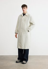 Νεαρός άντρας που στέκεται φορώντας μακρύ μπεζ παλτό, σκούρο παντελόνι με φαρδιά μπατζάκια και γκρι παπούτσια πάνω σε λευκό φόντο.