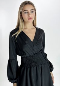 Zwarte jurk met een diepe V-hals, lange pofmouwen en een gesmokte taille. De stof heeft een gladde, satijnachtige textuur met een vloeiende rok.