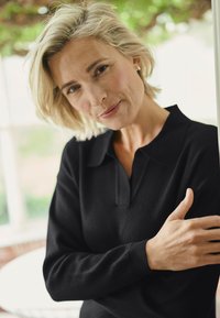 Black pullover with a collar, featuring textured fabric and long sleeves. The model has short, light blonde hair and is positioned against a light background.