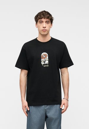 Nuori mies, jolla on kiharat ruskeat hiukset, musta T-paita, jossa oranssinvärinen kukkakuvio, ja vaaleansiniset farkut, seisoo yksinkertaista taustaa vasten.