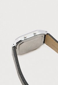 Sidabrinis kvadratinis skaitmeninis laikrodis su dviem šoniniais mygtukais ir juodu odiniu dirželiu, rodomas iš nugaros šviesiame fone.