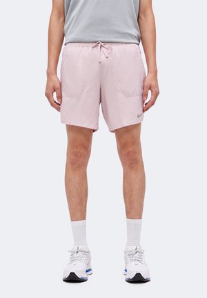 Persoană purtând pantaloni scurți roz pal cu șnur, tricou gri, șosete albe și pantofi sport albi, stând în fața unui fundal simplu.