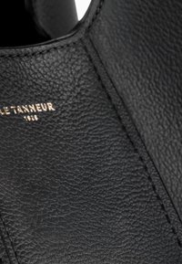 Borsa in pelle nera con superficie testurizzata, cuciture visibili e logo metallizzato color oro. Vista ravvicinata dei dettagli del design elegante.