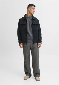 Schwarze Zip-Jacke über einem grauen Pullover, kombiniert mit grauen Hosen und beigen Slippern. Die Jacke hat zwei Brusttaschen.
