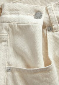 Denim tkanina kremaste boje sa suptilnim rebrima. Ima bočnu džepu i metalne dugmadi s ugraviranim detaljima.