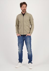 Chaqueta cremosa con cremallera y cuello alto, combinada con vaqueros azules. Zapatillas blancas completan el conjunto, sobre un fondo blanco liso.