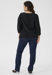 Zwarte textuur blouse met pofmouwen en knoop aan de achterkant van de nek, gecombineerd met donkerblauwe rechte jeans en zwarte hoge hakken.