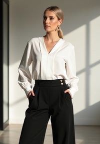 Biała, długa bluzka z rękawami i dekoltem w kształcie litery V, z elastycznymi mankietami, w połączeniu z czarnymi, szerokimi spodniami o wysokim stanie, z złotymi guzikami.