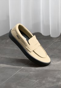 Beige slip-on cipő kerek orral, velúr anyagból és fekete gumi talppal. Jellemzője egy pánt a lábfejen, amely biztos illeszkedést biztosít.