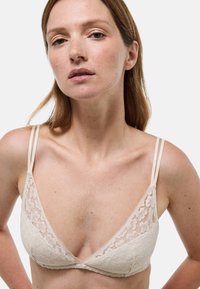 Brassière en dentelle de couleur beige clair avec de fines bretelles réglables, des bonnets triangulaires et un motif floral le long des bords. Texture douce et design minimaliste.