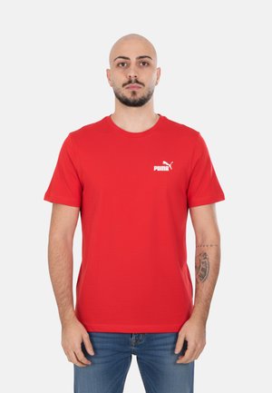 Uomo con testa calva e barba, che indossa una maglietta rossa Puma e jeans blu, in piedi contro uno sfondo bianco semplice.