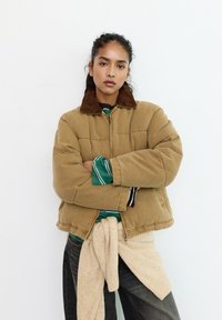 Ung kvinde står med korslagte arme iført en tan farvet dunjakke, grønstribet skjorte, sorte jeans og en beige sweater bundet om taljen.