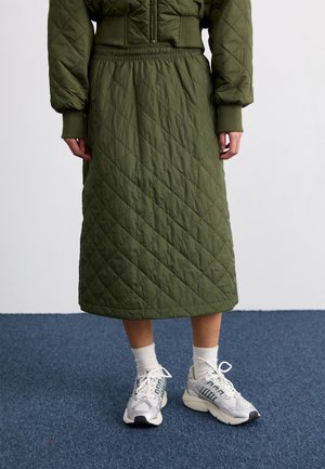 Personne portant une jupe midi matelassée vert olive et une veste assortie, avec des chaussettes blanches et des baskets blanches, debout sur un sol recouvert de moquette bleue.