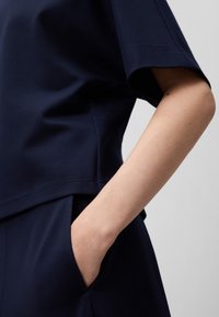 Haut en tissu doux de couleur bleu marine avec des manches courtes. Présente une coupe carrée et des poches sur les côtés. Détails de couture le long des manches et de l'ourlet.