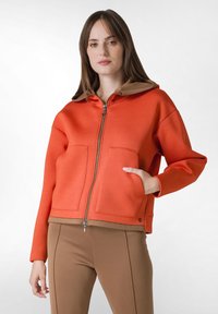 Orange zip-up hoodie med en brun huva, två framfickor och ribbad midja. Slät tygtextur, något oversized passform.