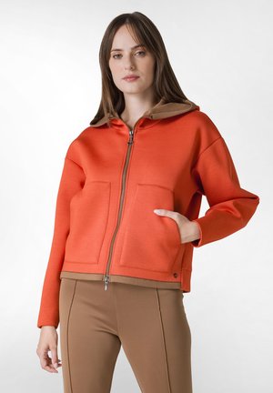 Orange zip-up hættejakke med en brun hætte, to forlommer og ribbet talje. Glat stofstruktur, let oversized pasform.