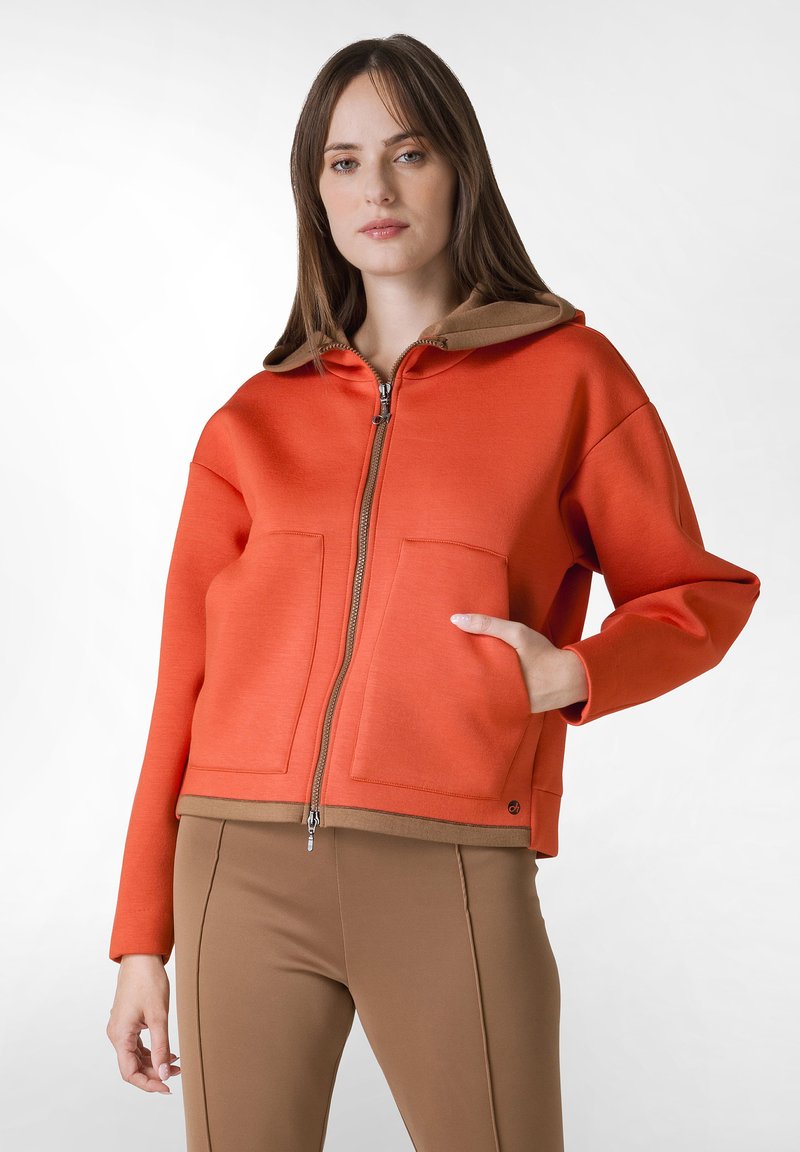 Orange zip-up hoodie med en brun huva, två framfickor och ribbad midja. Slät tygtextur, något oversized passform.