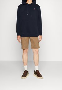 Marineblauwe hoodie met voorzak en trekkoorden, gecombineerd met beige shorts en donkerbruine sneakers met lichtgekleurde zolen en witte sokken.