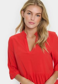 Blouse rouge avec un col à volants, manches trois quarts et un ourlet à étages. Tissu lisse avec un éclat subtil, design minimaliste.