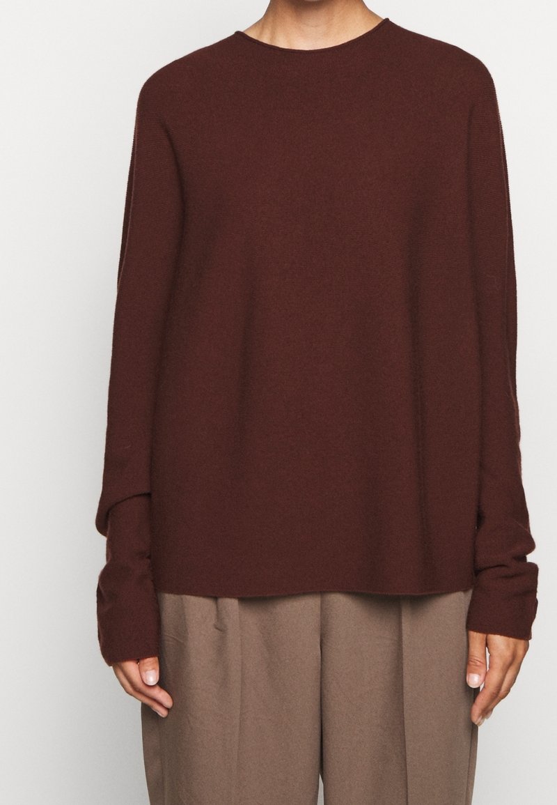 Personne portant un pull ample à manches longues marron foncé et un pantalon plissé marron clair, debout, les mains détendues le long du corps.