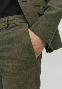 Zöld vászon öltöny finom textúrával; közelkép a nadrágzsebről, amelyben egy kéz pihen. Testre szabott fazon, tiszta vonalak, minimalista részletezés.