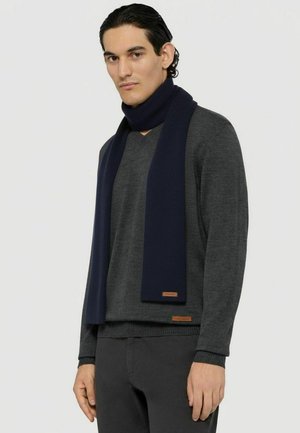 Marineblåt ribstrikket tørklæde i strikket materiale, båret over en mørkegrå V-hals sweater. Har et lille brunt lædermærke nederst.