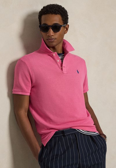 Άνδρας με μαύρα γυαλιά ηλίου, ροζ πόλο πουκάμισο με ανεβασμένο γιακά πάνω από ριγέ πουκάμισο και μπλε παντελόνια με λεπτές ρίγες, γέρνει σε έναν τοίχο.