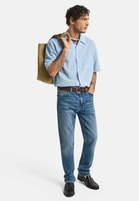 Camisetta azzurra a maniche corte, con taschino, abbinata a jeans blu, cintura marrone e scarpe nere. Borsa a tracolla compatta in olivo portata sulla spalla.