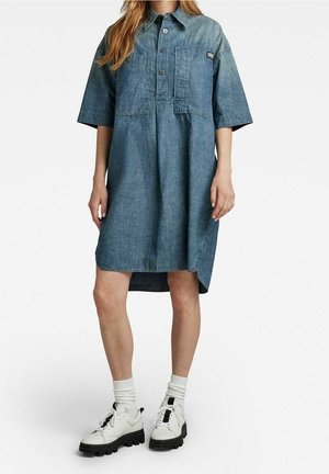 Kvinde iført en knælange blå denimkjole med korte ærmer, forlommer, hvide sokker og hvide sneakers med tyk sål.