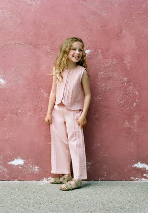 Jeune fille aux longs cheveux bouclés portant un haut rose sans manches, un pantalon assorti et des sandales vertes, debout contre un mur rose texturé.