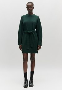 Gestricktes dunkelgrünes Kleid mit rundem Halsausschnitt, langen Ärmeln, Gürtel in der Taille und gerippten Säumen, kombiniert mit schwarzen Socken und Schuhen.