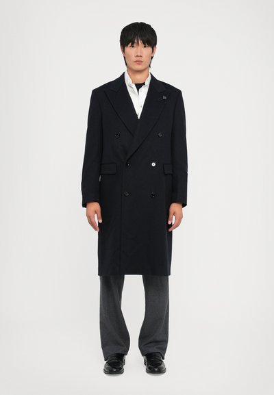 Cappotto navy doppiopetto con ampi risvolti, chiusura con bottoni e due tasche laterali, indossato su una camicia bianca e pantaloni grigi. Scarpe nere.