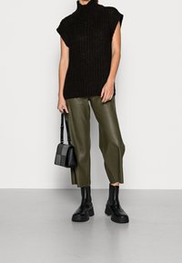 Czarny dziany bezrękawnik z golfem, oliwkowo-zielone szerokie spodnie z imitacji skóry, czarne botki oraz czarno-szara torebka na pasku przewieszanym przez ramię.