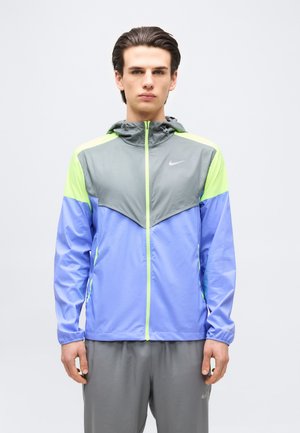 Junger Mann trägt eine farblich abgesetzte Kapuzen-Windjacke in Grau, Hellgrün und Blau, steht vor weißem Hintergrund.