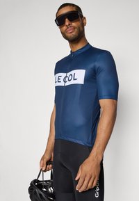Maillot de cyclisme bleu marine avec une fermeture éclair à l'avant, comportant un grand logo blanc "LE COL". Le modèle tient un casque noir et porte un short rembourré noir.