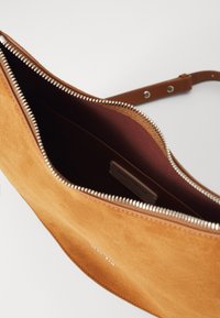 Borsa in suede beige con tracolla in pelle marrone e zip argentata. L'interno è di un marrone più scuro, con un'etichetta del brand ben visibile all'interno.