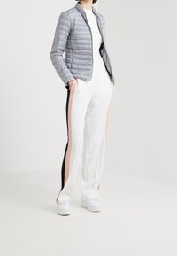 Ljusgrå pufferjacka med dragkedja framtill och horisontell quiltning, tillsammans med vita byxor med svarta och rosa sidostråk och knappar.