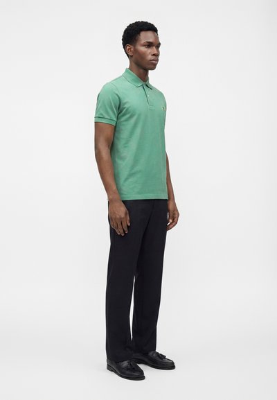 Homme debout portant un polo vert à manches courtes, un pantalon noir et des mocassins noirs à pampilles sur fond blanc uni.