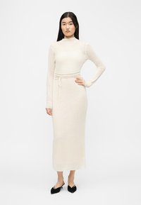 Une femme se tient debout, vêtue d'une robe crème en maille côtelée à manches longues et longueur cheville, avec une ceinture à la taille et des escarpins noirs à bout pointu et talons bas.