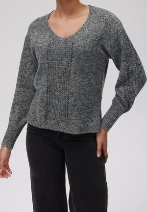 Personne portant un pull en maille gris à col en V avec un panneau texturé et des manches longues, associé à un pantalon noir sur un fond uni.