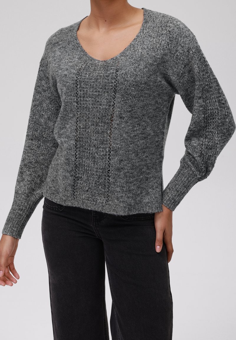 Personne portant un pull en maille gris à col en V avec un panneau texturé et des manches longues, associé à un pantalon noir sur un fond uni.