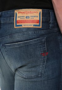 Vyblednuté modré riflové nohavice s viditeľnou koženou značkovou záplatou s nápisom "Diesel Industry" a červeným vyšitým logom na zadnom vrecku.