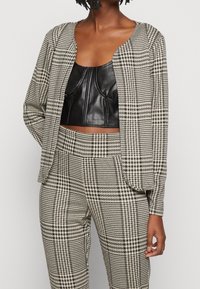 Veste courte à manches longues à motif pied-de-poule noir et blanc, associée à un haut ajusté en similicuir noir et un pantalon assorti.