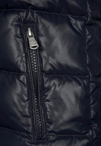 Námořnická puffy bunda s kapsou na zip. Materiál je lesklý a prošívaný, s horizontálním švem pro izolaci.