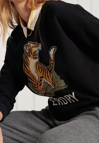 Svart sweatshirt med en stor broderad tigerdesign. Kragen har en gul och vit randig detalj. Texten "DEAD DRY" finns under tigern.