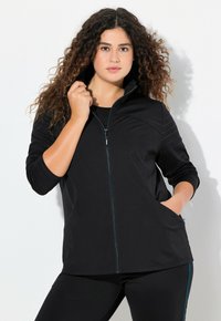 Svart zip-up jacka med hög krage, med texturerade axeldetaljer och sidofickor. Bärs över en svart topp, kombinerad med svarta leggings.