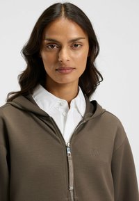 Khaki zip-up hoodie med en subtil logotyp, med ribbad nacke, mjuk textur och justerbar dragsko på huvan, lager på lager över en vit skjorta med knappar.
