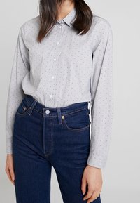 Camicia button-up grigia chiara con piccoli pois neri; maniche lunghe e colletto classico. Abbinata a jeans blu scuro a vita alta.