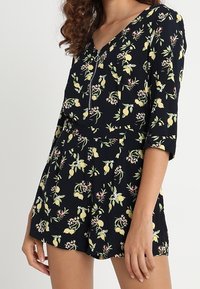 Svart romper med citron- och blommönster, dragkedja fram och trekvartsärmar, bärs av en person med lockigt hår.