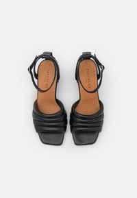 Mustat korot sandaalit neliömäisellä varpaalla, poimutetusta nahasta valmistettu yläosa ja nilkkasuljin. Sisäosassa on vaalea pohja, jossa on kohokuvioitu "PAVEMENT".